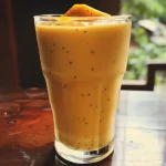 A vibrant, close-up view of a creamy mango smoothie in a glass with tropical fruit around it.