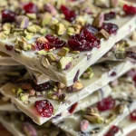 ['Close-up of colorful Cranberry Pistachio White Chocolate Bark with nuts and dried fruits.', 'Side view of white chocolate bark topped with green pistachios and red cranberries.', 'Detailed shot of Cranberry Pistachio White Chocolate Bark showing texture and ingredients.', 'A vibrant piece of bark featuring white chocolate, pistachios, and cranberries.']