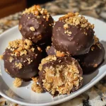 ['Close-up side view of crispy peanut butter toffee truffles with a glossy chocolate coating.', 'Detailed image of round truffles featuring a crunchy exterior made from peanut butter and toffee bits.', 'Side view of several peanut butter toffee truffles sprinkled with toffee bits on a white plate.', 'Irresistible shiny milk chocolate toffee truffles with a crispy texture, highlighted in natural light.']