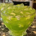 ['A close-up view of a vibrant green drink in a glass, decorated with ice and fruit.', 'A side view of a refreshing, green punch served in a glass with ice cubes and a colorful garnish.', 'A detailed look at a green beverage in a glass, featuring bubbles and a slice of pineapple on the rim.', 'A juicy green punch with visible ice and a bright color, captured from the side in natural lighting.']