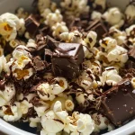 ['Close-up of popcorn drizzled with dark chocolate and sprinkled with sea salt.', 'Side view of salted dark chocolate popcorn in a bowl, glistening with chocolate.', 'Handful of popcorn coated in dark chocolate, dusted with flaky salt.', 'Textured view of dark chocolate popcorn with visible salt crystals on top.']