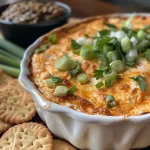 ['Close-up view of Tombstone Dip in a bowl with garnishes.', 'Side view of a creamy dip made with cream cheese, cheddar, and garnished with scallions.', 'Juicy Tombstone Dip displayed in a white bowl, surrounded by Town House Crackers.', 'Detailed shot of Tombstone Dip with pimenton peppers and black olive tapenade.']
