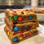 ['Close-up view of golden-brown chocolate chip M&M blondies with colorful M&M pieces.', 'Side view of freshly baked blondies topped with chocolate chips and M&Ms.', 'Juicy chocolate chip M&M blondies displayed on a plate, highlighting their chewy texture.', 'Delicious blondies featuring chunks of M&Ms and chocolate chips, captured in rich detail.']